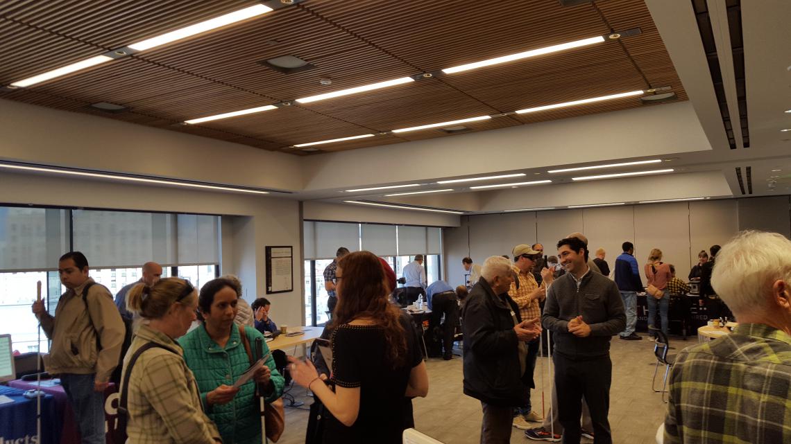 Picture of the public space room at the Lighthouse for the Blind in San Francisco with people checking out devices and talking with other attendees.